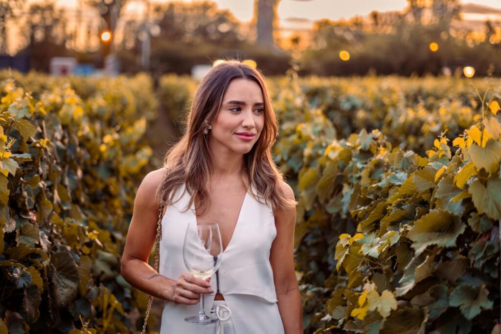 A woman with a wine glass walks through a lush vineyard at sunset, enjoying an evening outdoors