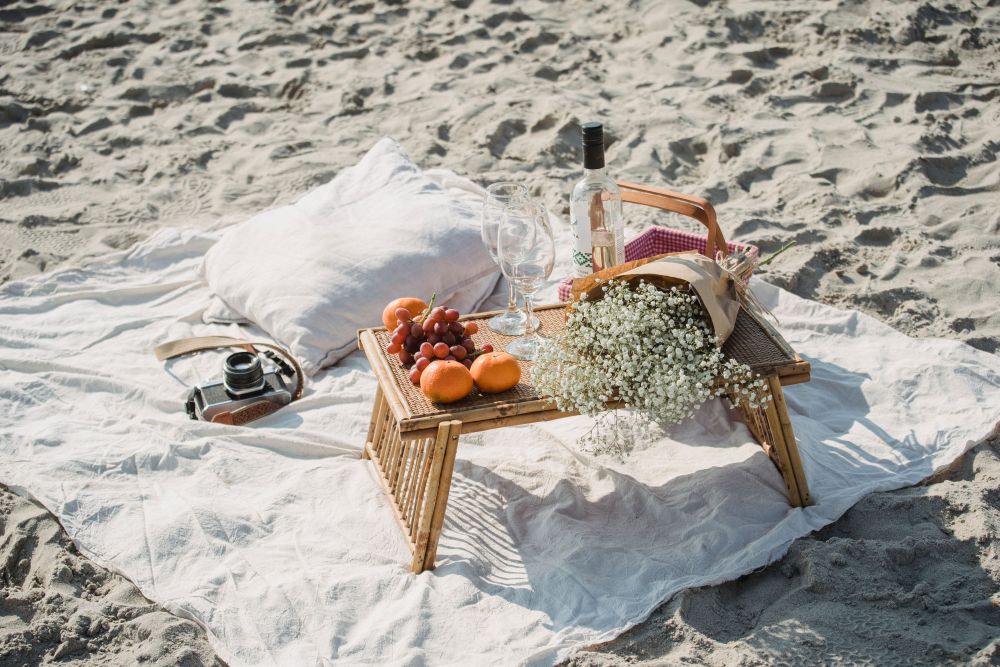 A beach picnic laid out on a blanket with cushions, food and drinks by the sea